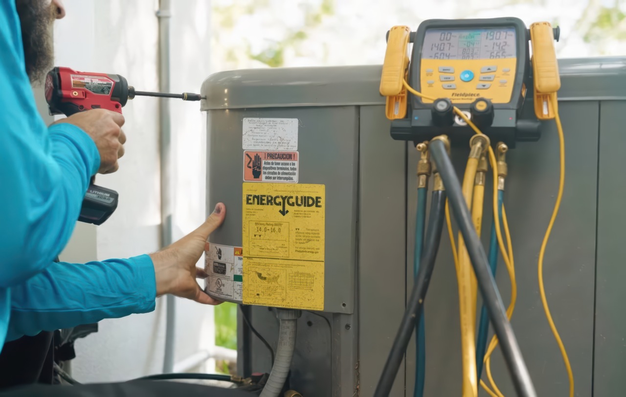 A bearded technician in a teal shirt operates a red Milwaukee cordless drill on a gray outdoor AC unit, with a yellow EnergyGuide sticker rated 14.0-16.0 visible on the panel and a Fieldpiece digital manifold gauge with colored refrigerant hoses connected on the right.