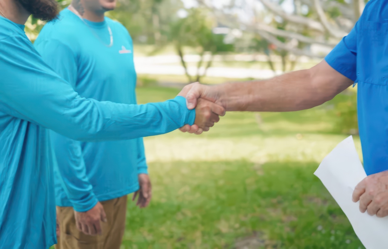 Close-up of an AC technician in teal uniform shaking hands with a customer holding paperwork in a residential yard.