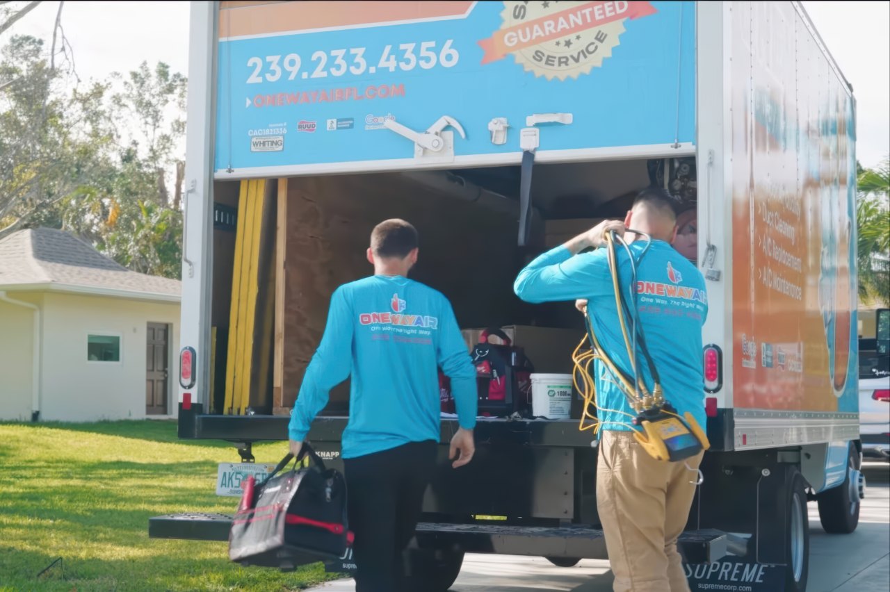 Two service workers in One Way Air uniforms retrieve equipment from a branded truck parked near a residential home under bright daylight.