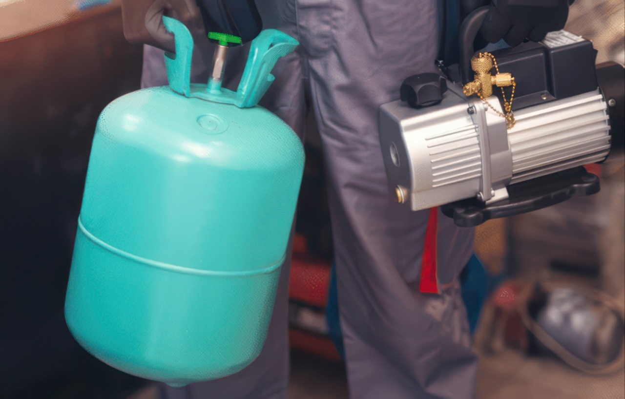 An HVAC technician wearing a gray shirt holds a turquoise refrigerant tank in one hand and a silver vacuum pump with brass valve connections in the other hand.