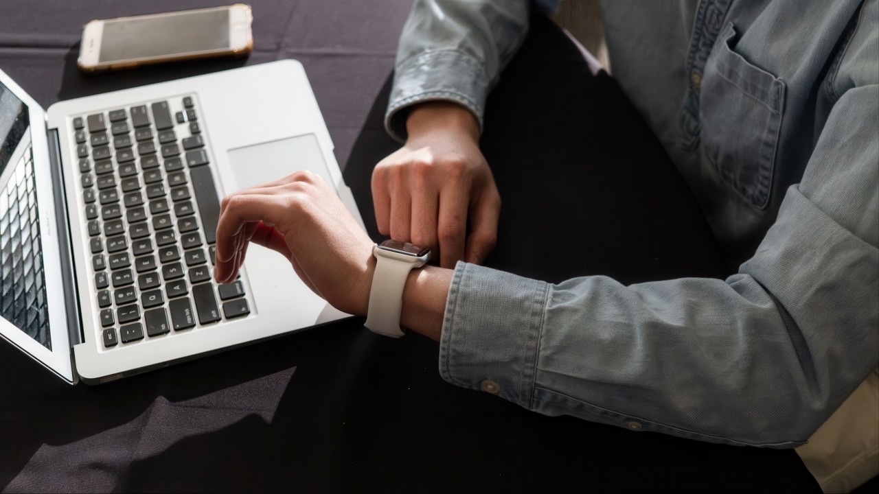 Standard
Person in gray shirt checks white smartwatch on wrist while sitting with open laptop, smartphone, and wire basket on dark desk