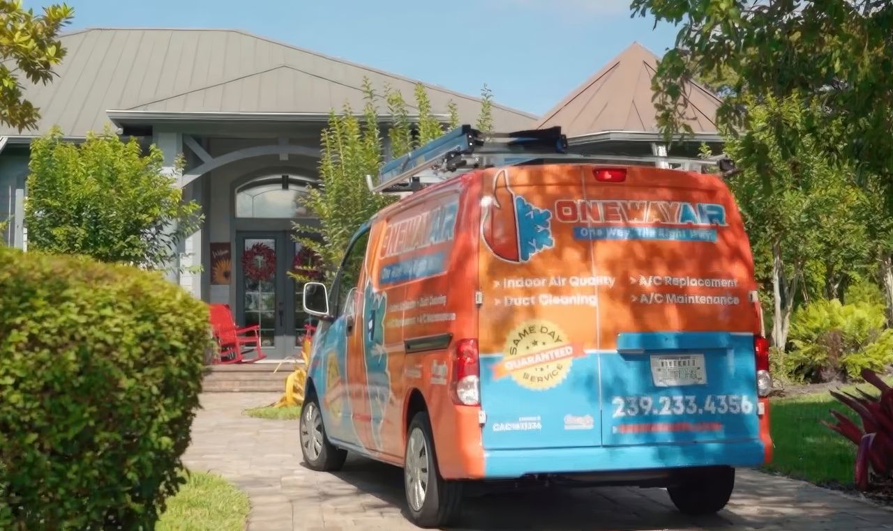 An orange and blue One Way Air service van equipped with roof ladders parks on a paved driveway in front of a house.