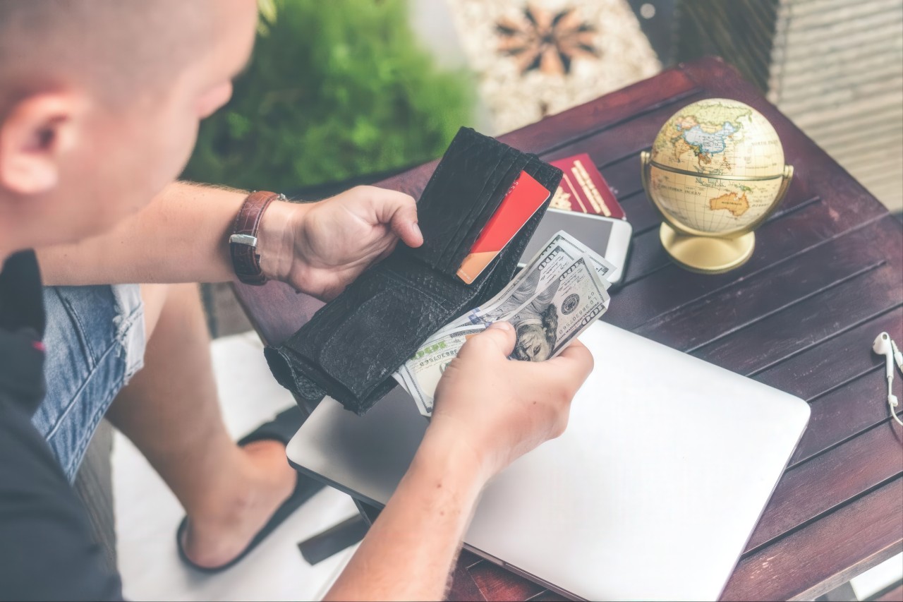 A man with a brown leather watch removes cash from a black wallet containing dollar bills, credit cards, and red passports while seated at a dark wooden desk.