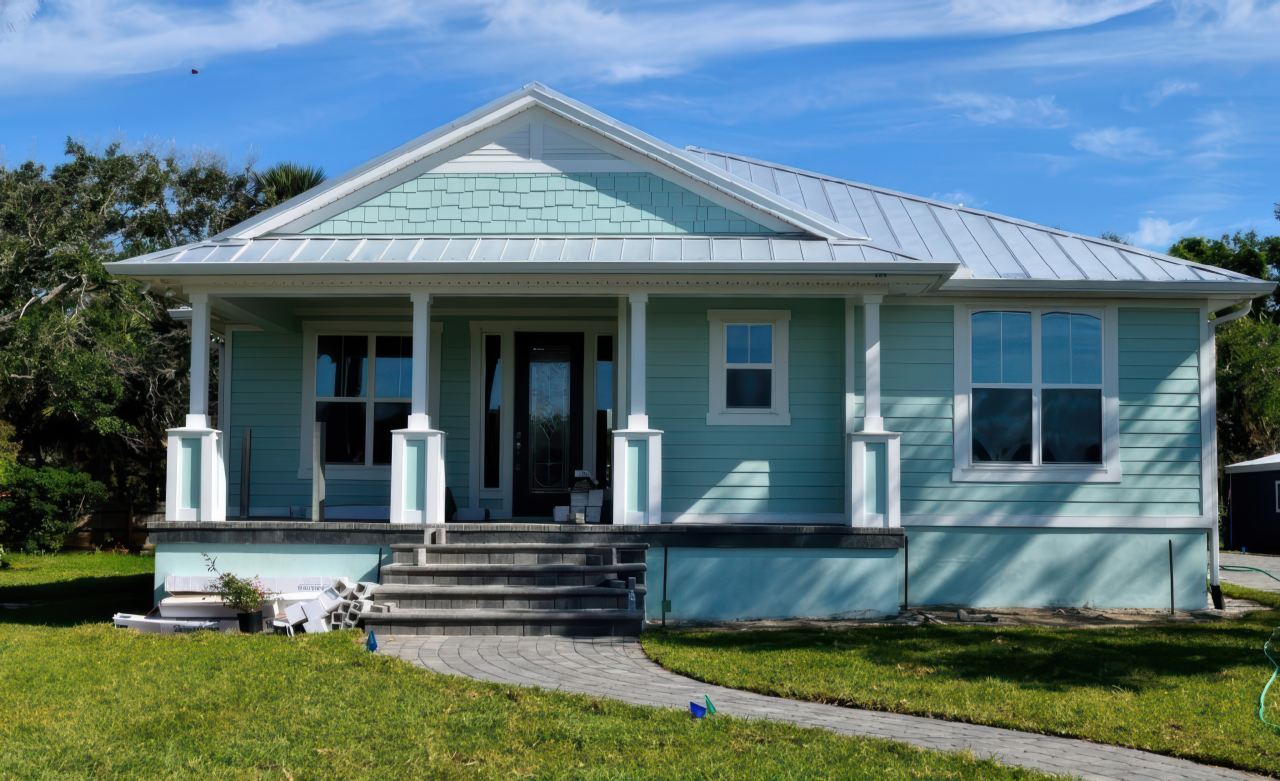Front view of a pastel blue residential home featuring a raised porch, white columns, large windows, and a curved paved path.