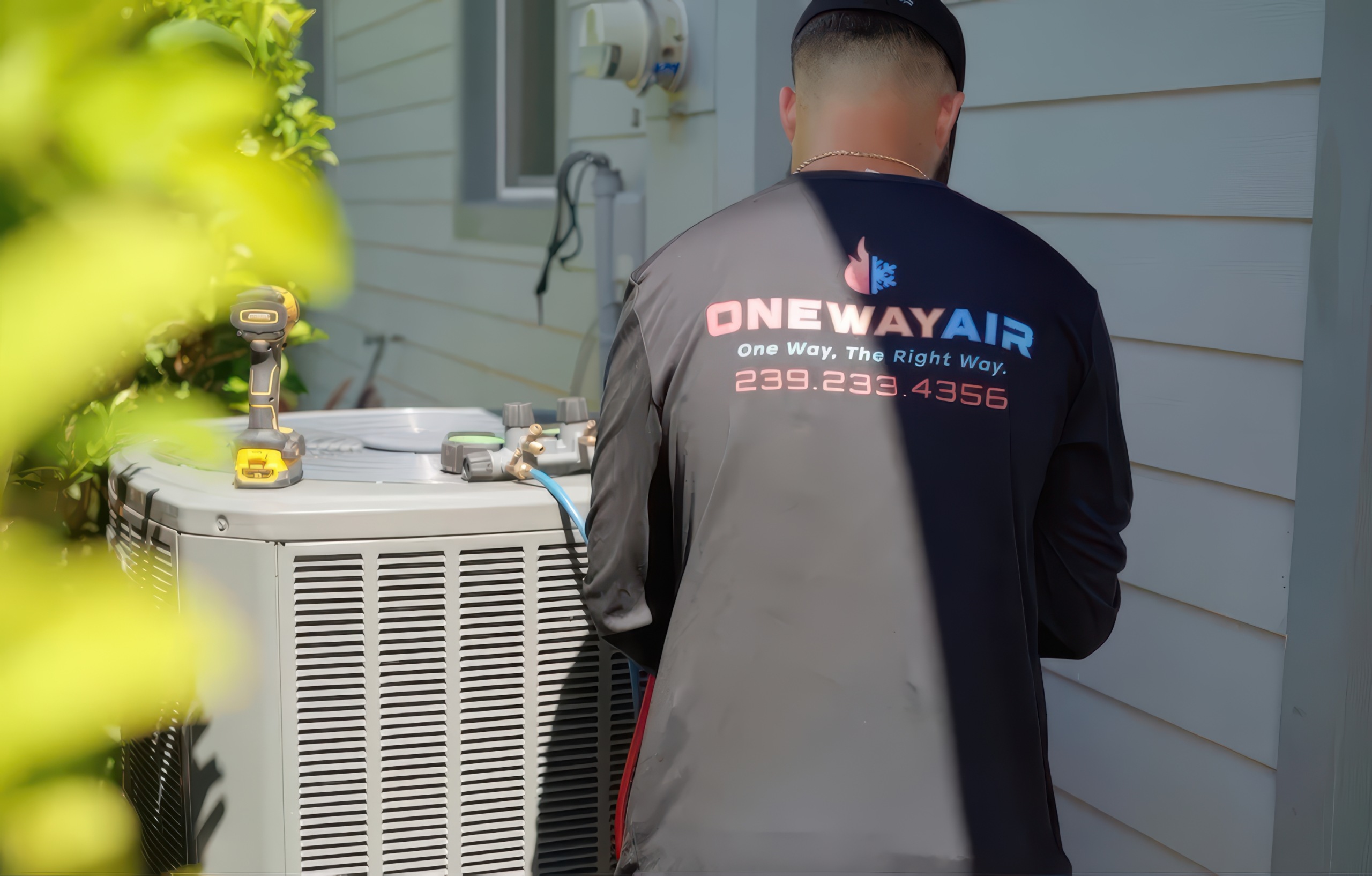 HVAC technician in branded work shirt services gray outdoor AC condenser beside white house with yellow power drill resting on unit top.