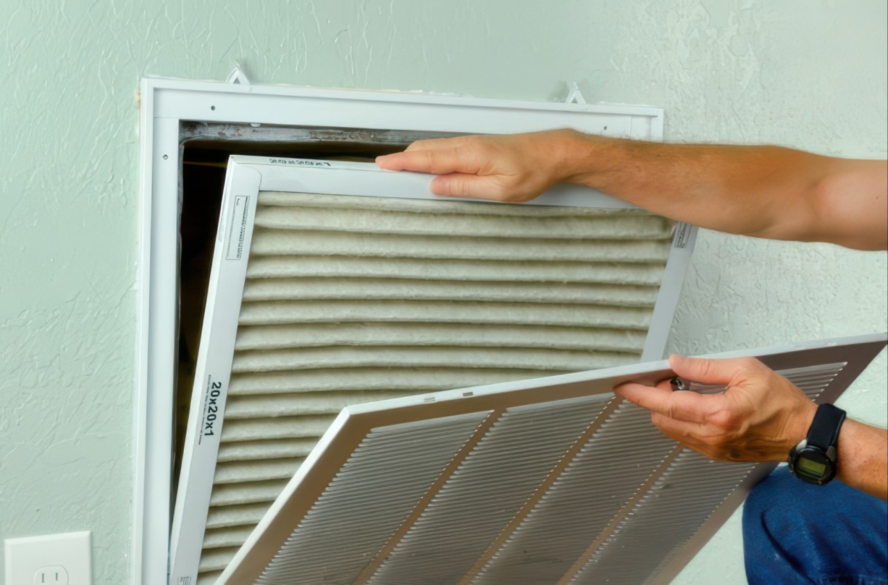 A man removes a beige pleated air filter from a white rectangular vent cover mounted on a light green wall. The dirty filter shows visible dust buildup while the dark vent opening is exposed behind the cover.