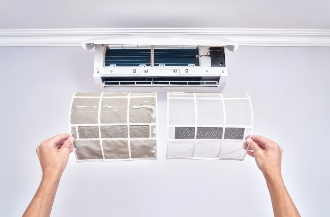 Two hands hold a pair of air filters for comparison below an open white wall-mounted air conditioner on a light blue wall. The left filter shows heavy gray dust accumulation while the right filter appears clean and white, demonstrating the difference between dirty and new filters.