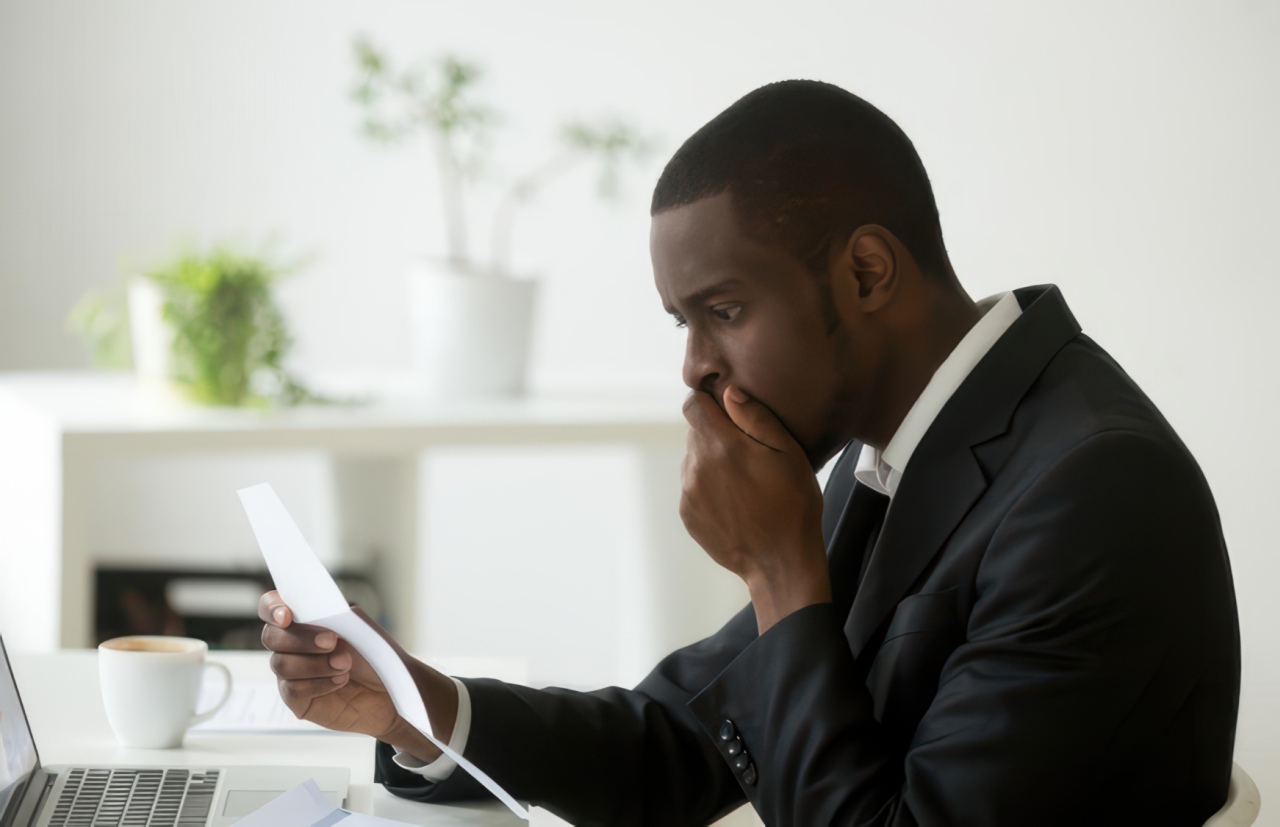 A man wearing a black suit and white shirt sits at a white desk reading a paper document with a worried expression, his hand covering his mouth.
