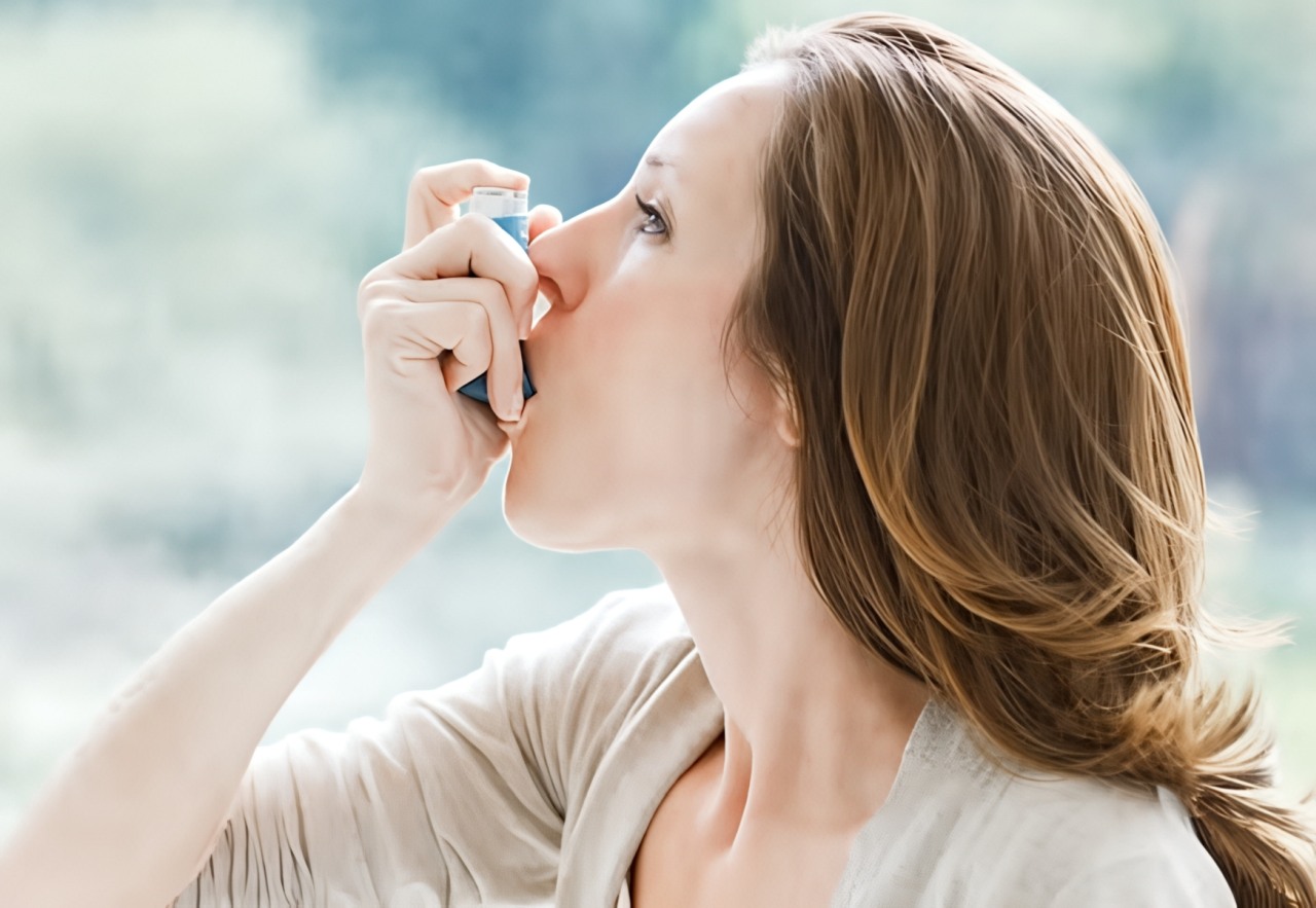 Woman with brown hair wearing beige shirt uses blue and white asthma inhaler against blurred outdoor background.