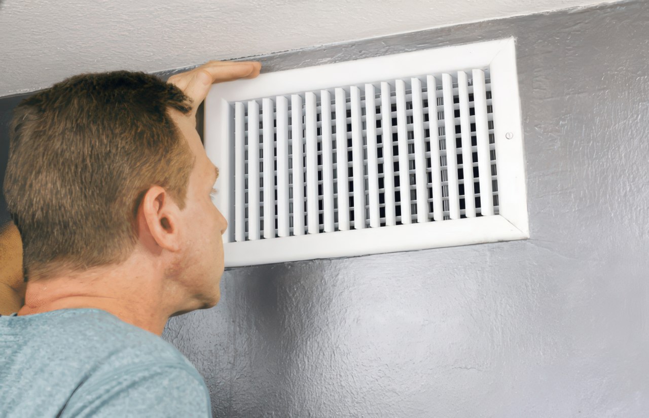 This photograph shows a focused middle-aged man inspecting a white air vent on a grey wall. He rests his hand on the frame while looking through the slats in a quiet indoor setting.