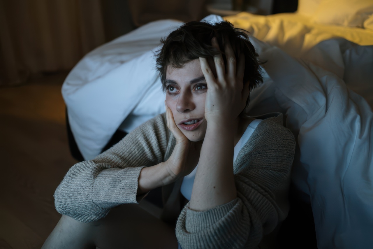 A young woman with short hair leans against a bed while holding her hands to her face in this dimly lit room photograph.