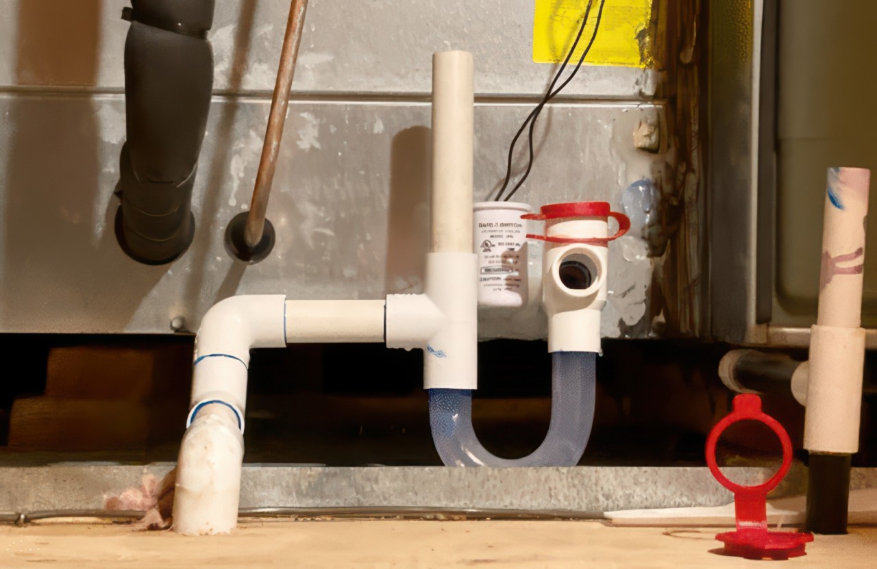 A technical photograph shows white PVC pipes and a translucent U-trap connected to a metallic furnace in a dimly lit utility space.