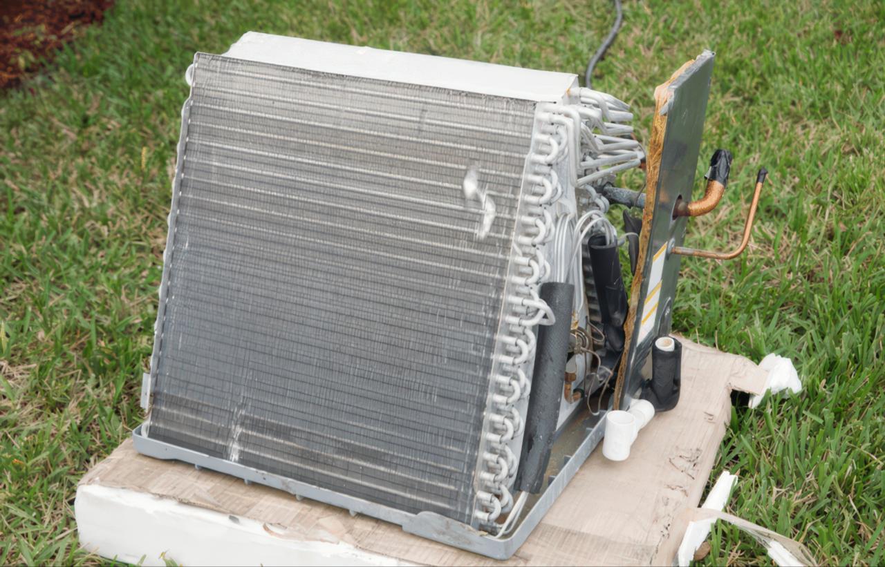 This photograph shows a large cooling coil with metallic fins and copper tubing sitting on a cardboard sheet. The object is positioned on a grassy surface under natural light to highlight the design of the HVAC component.