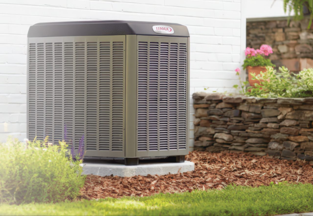 Tan and gray air conditioning condenser sits on white concrete base surrounded by brown mulch, green lawn, and stone retaining wall.