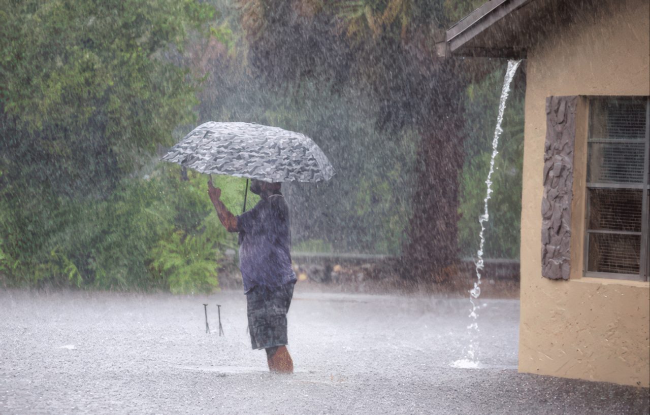 Man holding patterned umbrella stands in intense rainfall as water pours from roof gutter near a beige residential building