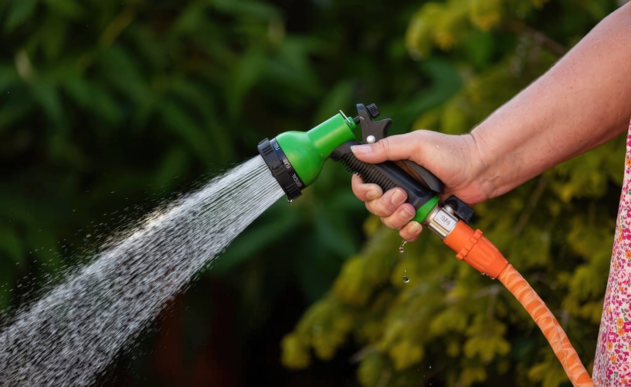 Hand grips green and black garden hose sprayer nozzle with orange hose attached, spraying water against green foliage background.
