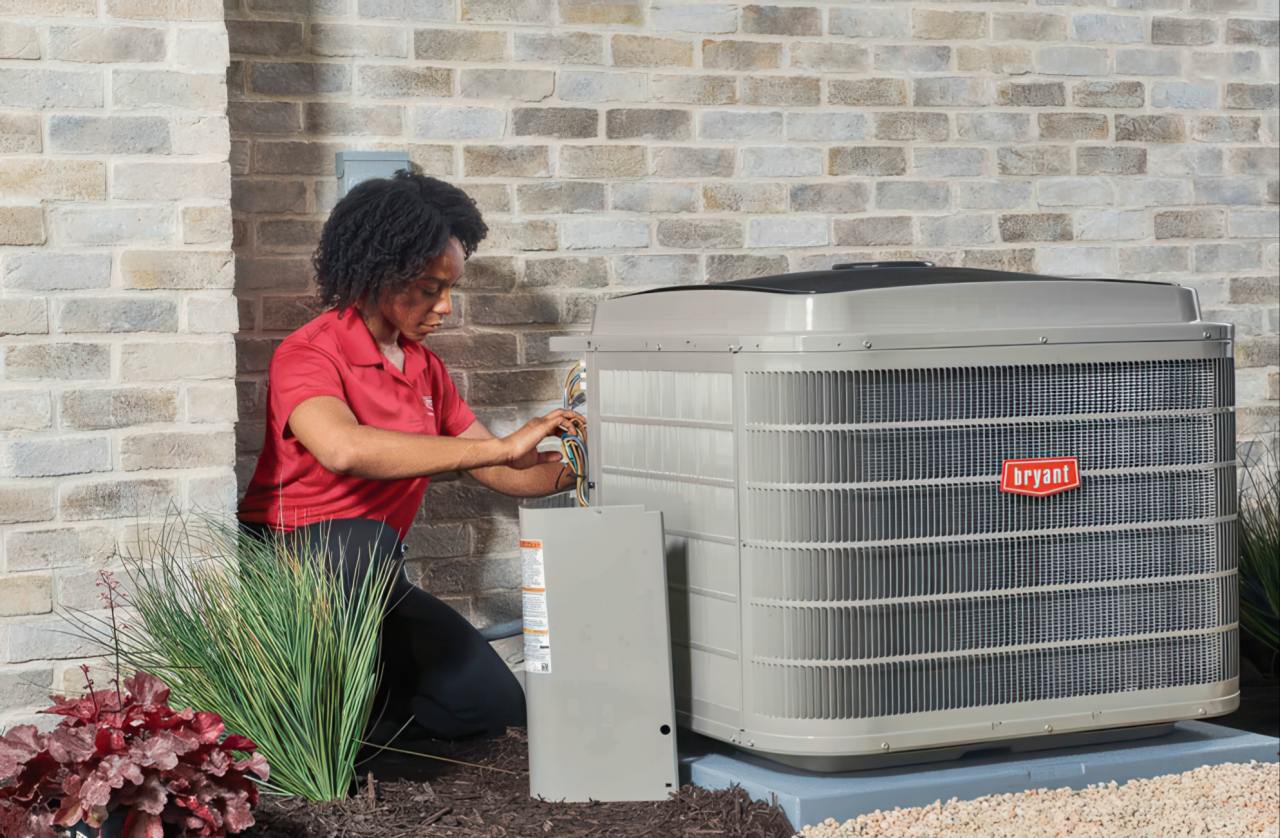 Woman wearing red polo shirt performs maintenance on gray outdoor air conditioning condenser unit positioned against tan brick residential wall.