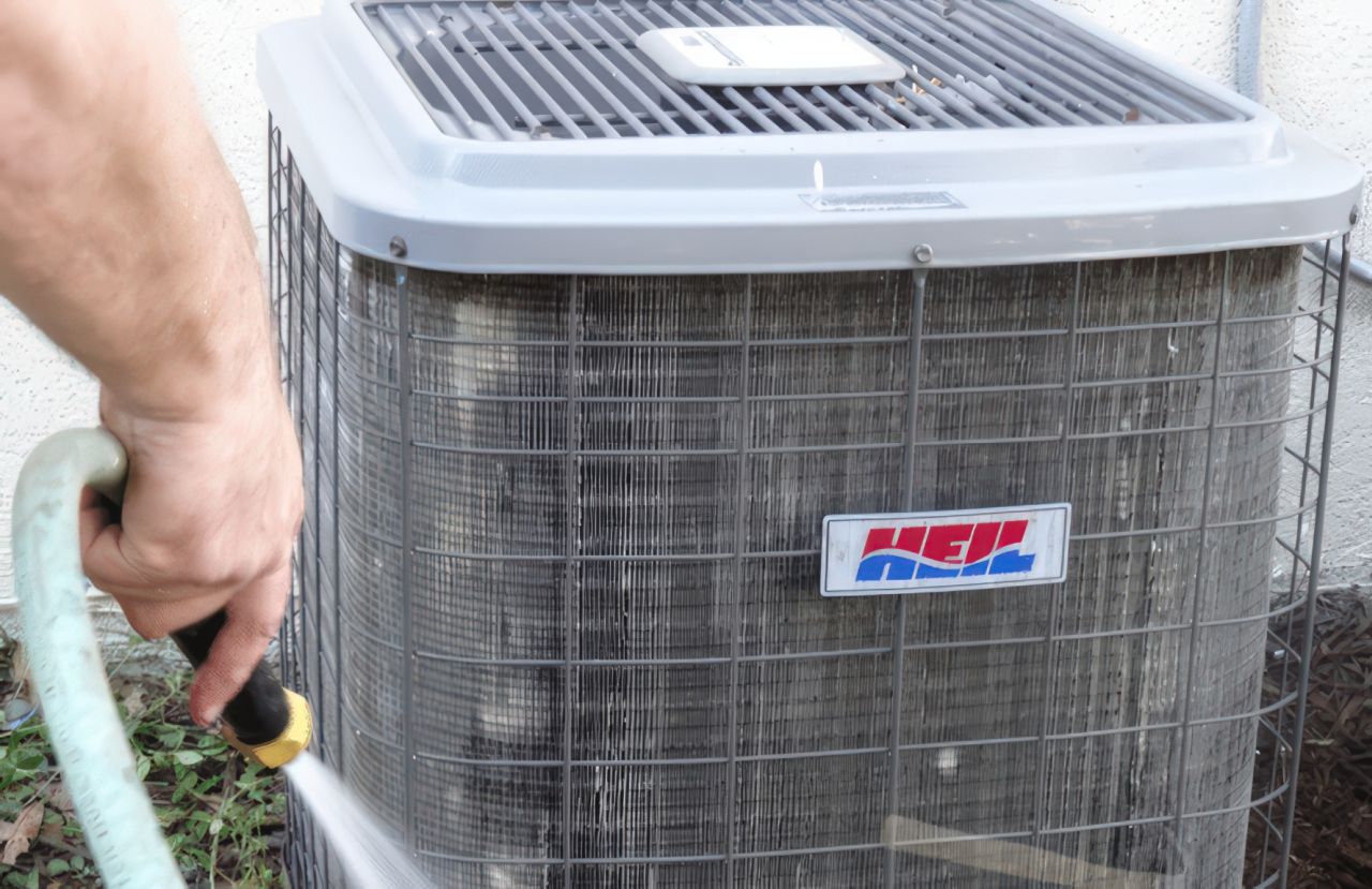 A hand grips a green garden hose while spraying water onto a gray outdoor air conditioning unit with a dirty coil, illustrating can i spray my ac unit with water while running. The cooling equipment sits against a white exterior wall with ground vegetation visible below.