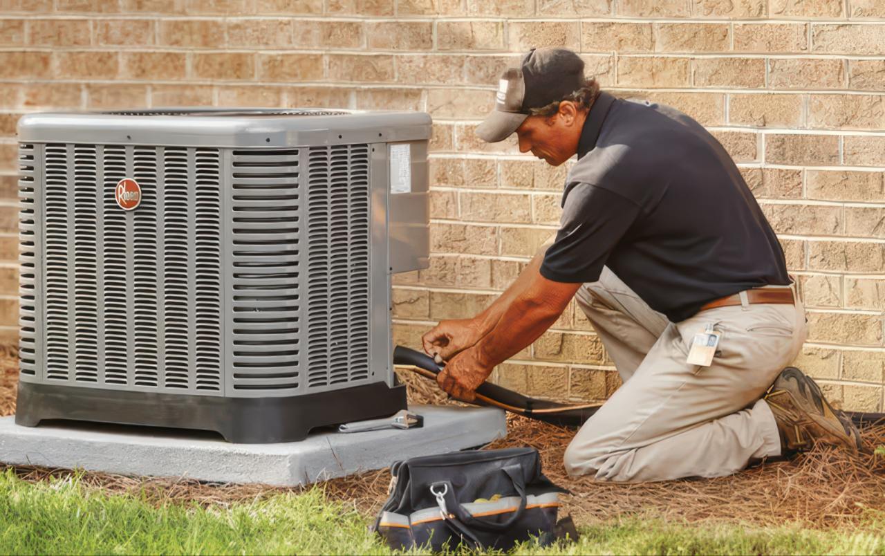 This professional photograph shows a middle-aged technician kneeling on pine straw to service a Rheem AC unit. He wears a black shirt and brown cap while a tool bag sits on the ground nearby.