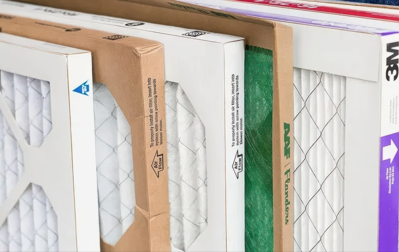 Various brands of air filters are stacked together, featuring both pleated white material and a green fiberglass mesh. Clear labels on the white and tan frames provide guidance on proper air flow direction.