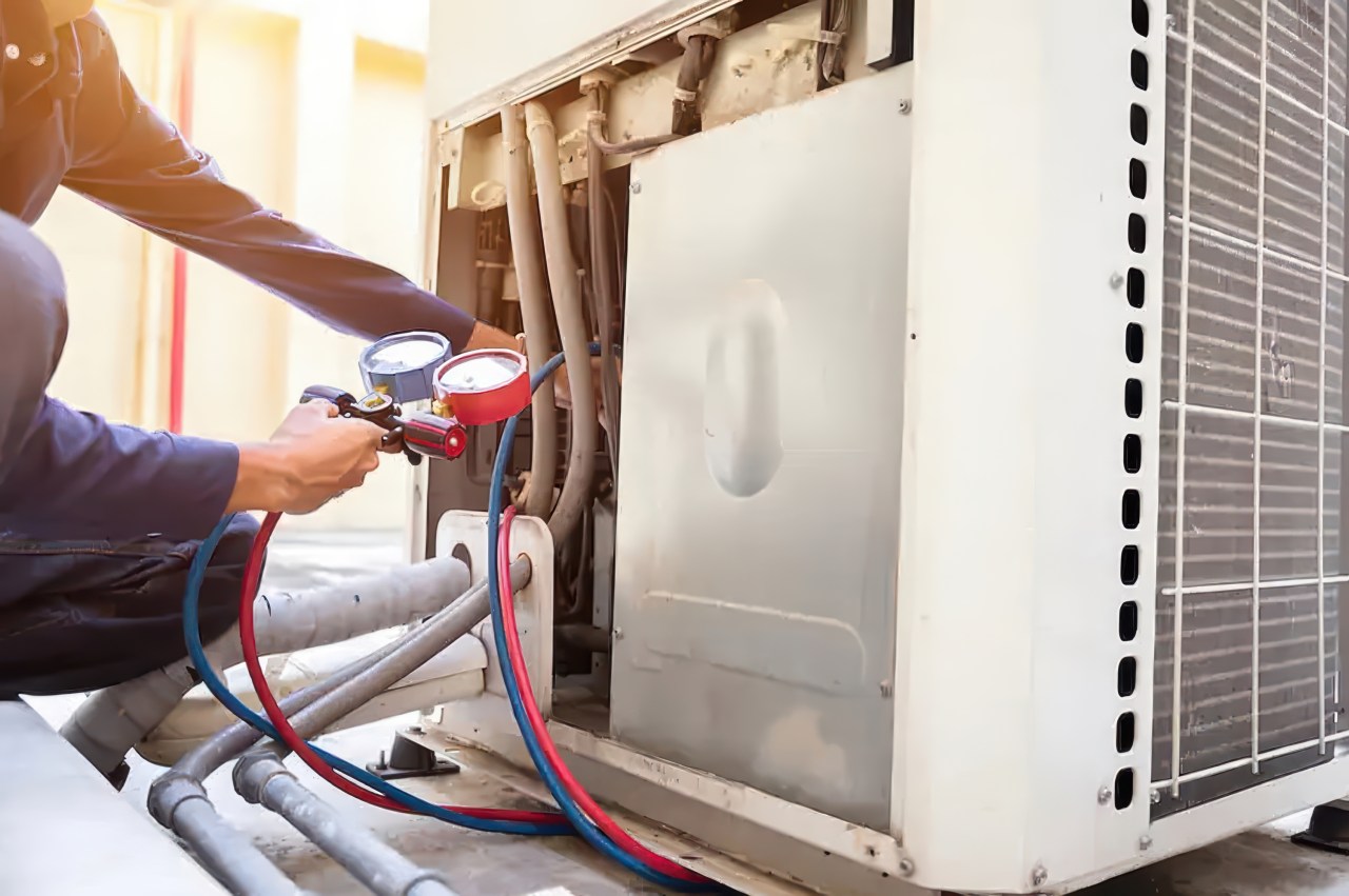 A middle-aged man kneels down on a rooftop, using a manifold gauge set with red and blue gauges and hoses to service an open outdoor air conditioning unit. Bright sunlight illuminates the scene, showing exposed components and background building walls.