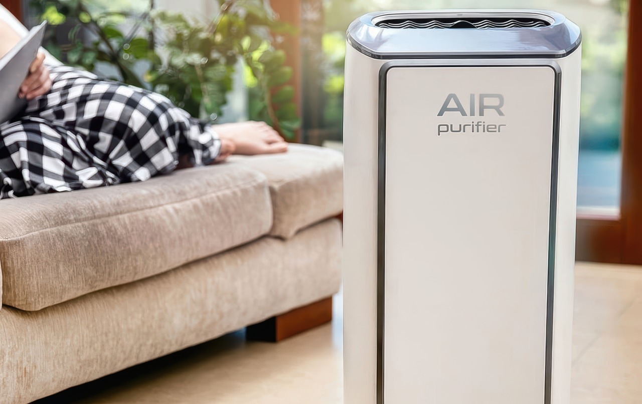 This photograph features a tall white air purifier with grey trim placed in a bright living room near a young person lounging on a tan sofa. Green plants and a large window create a calm and peaceful atmosphere in the background.