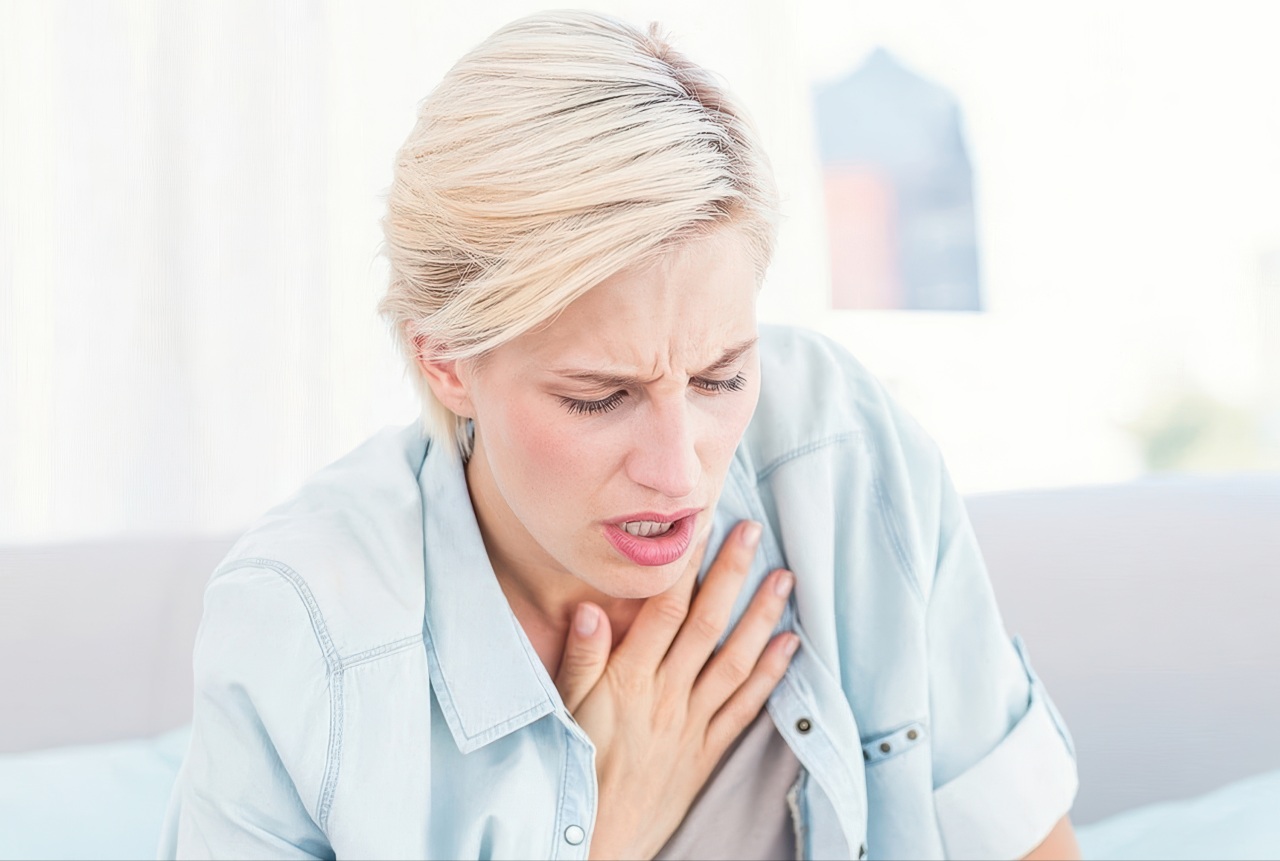 This photograph shows a young woman wearing a light blue shirt who appears to be in pain or having trouble breathing. She sits with a pained expression and holds one hand over her chest against a soft white background.