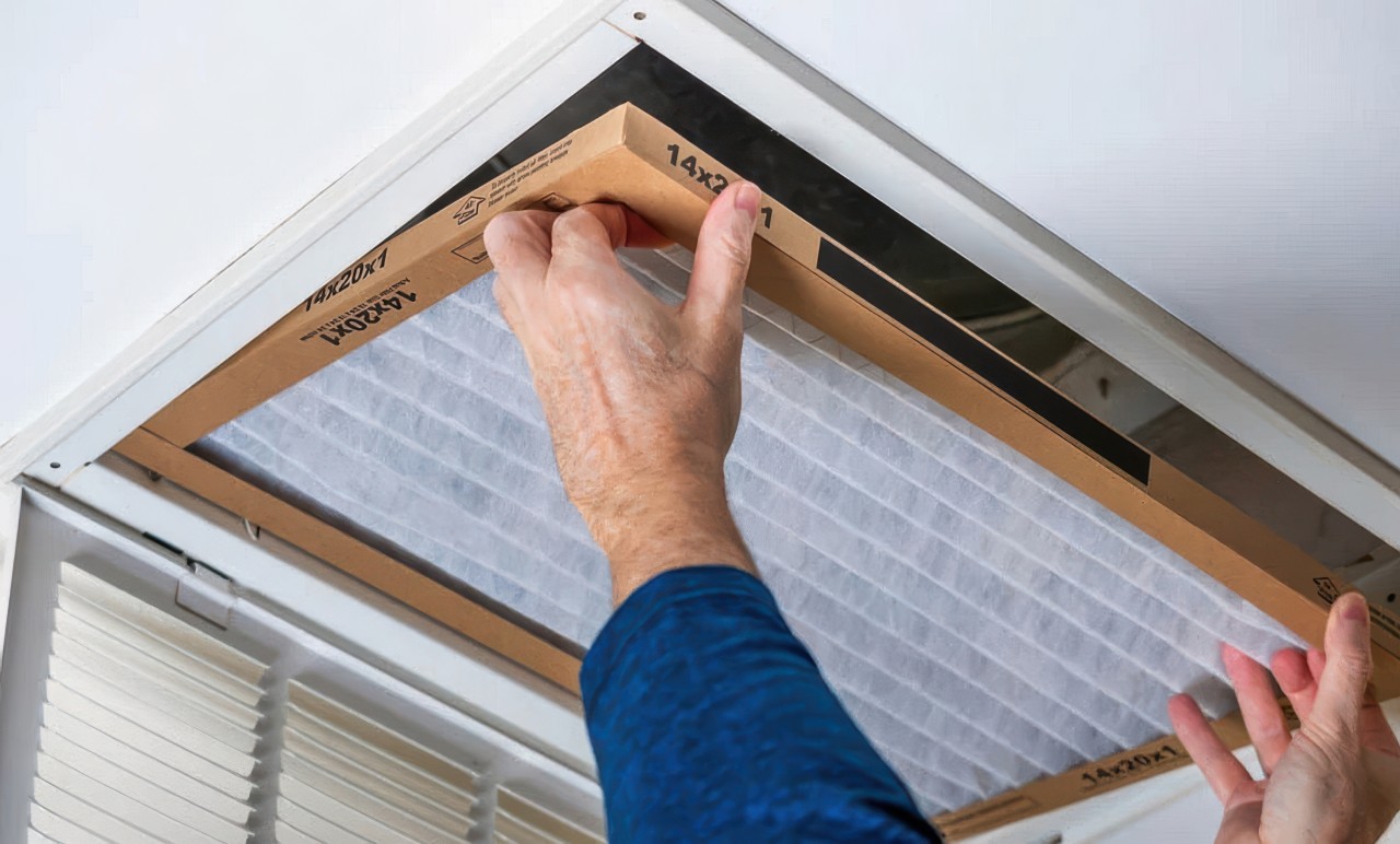 This photograph shows a person in a blue shirt replacing a white pleated air filter into a ceiling vent. The tan cardboard frame of the filter clearly displays its dimensions of 14 by 20 by 1.