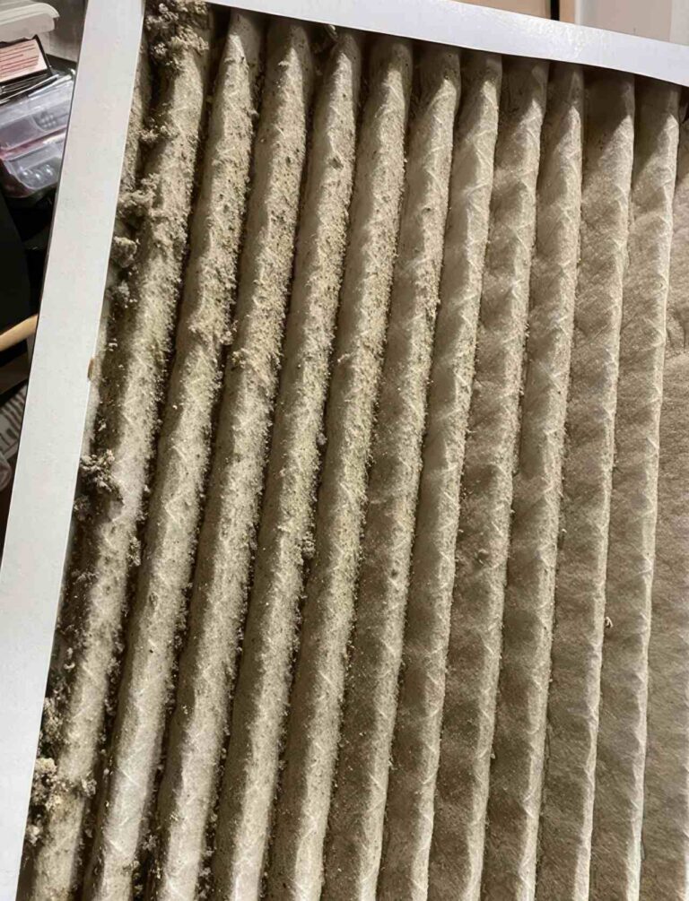Vertical ridges of an HVAC filter are heavily clogged with a thick layer of grey and tan dust. The white outer frame borders the dirty filtration material in this close up photograph.