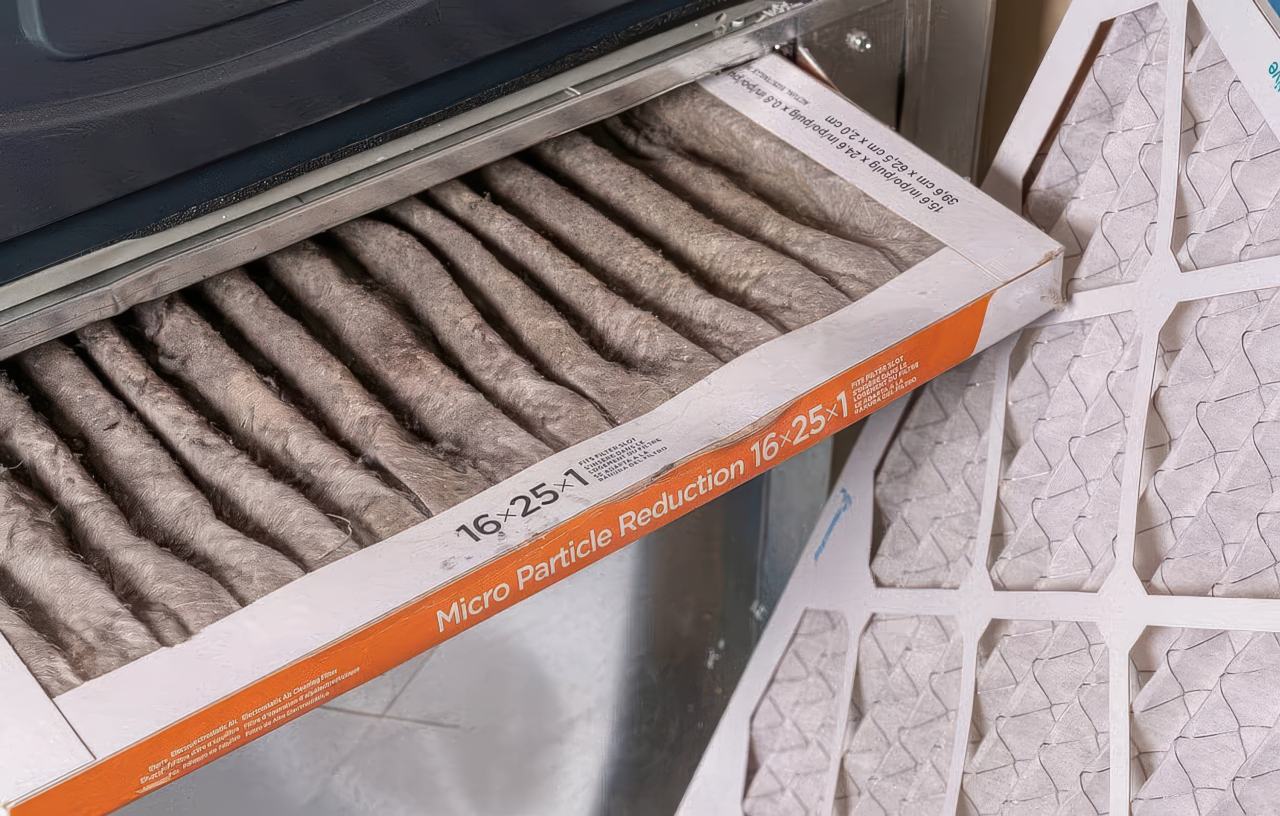 This photograph captures the contrast between a clogged air filter covered in thick grey dust and a pristine white replacement. The used filter is partially inserted into a metal HVAC slot and shows an orange border with printed dimensions.
