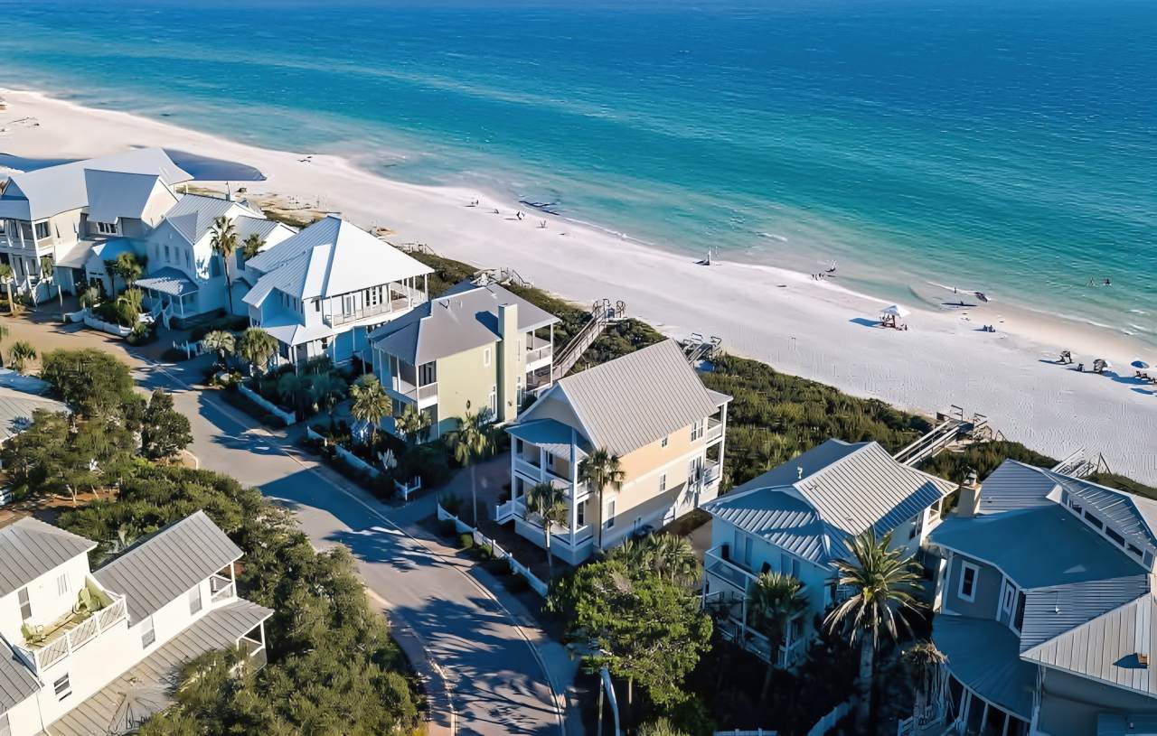 An aerial view captures beachfront homes in Florida with gray and white metal roofs nestled among palm trees and green vegetation along a white sandy beach.