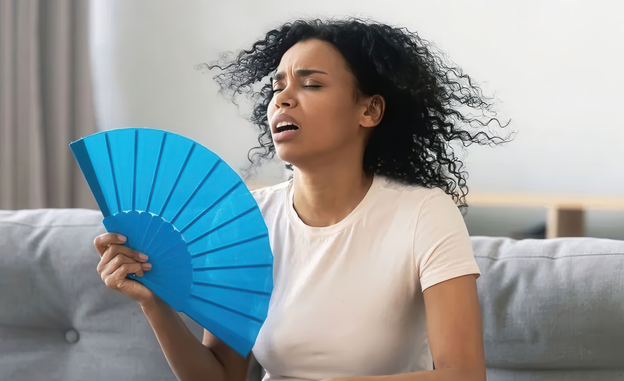 A woman with black curly hair wearing a beige shirt fans herself with a bright blue folded paper fan while sitting indoors. She appears overheated with her mouth open and hair blowing as she seeks relief in a living room with a gray couch and curtains visible behind her.