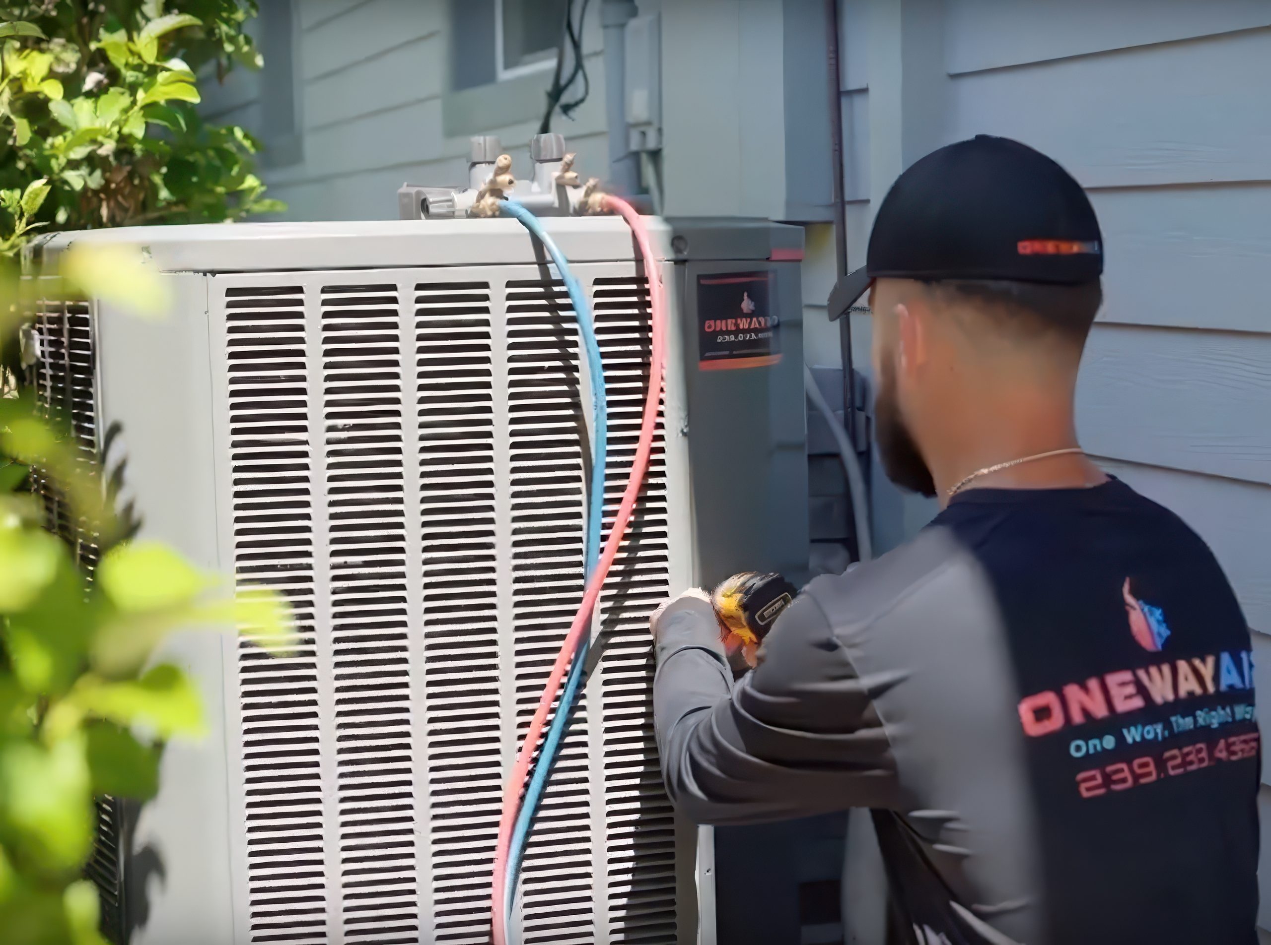 HVAC technician in a black ONEWAY AIR uniform and cap servicing an outdoor air conditioning unit using a power drill, with red and blue refrigerant hoses attached.