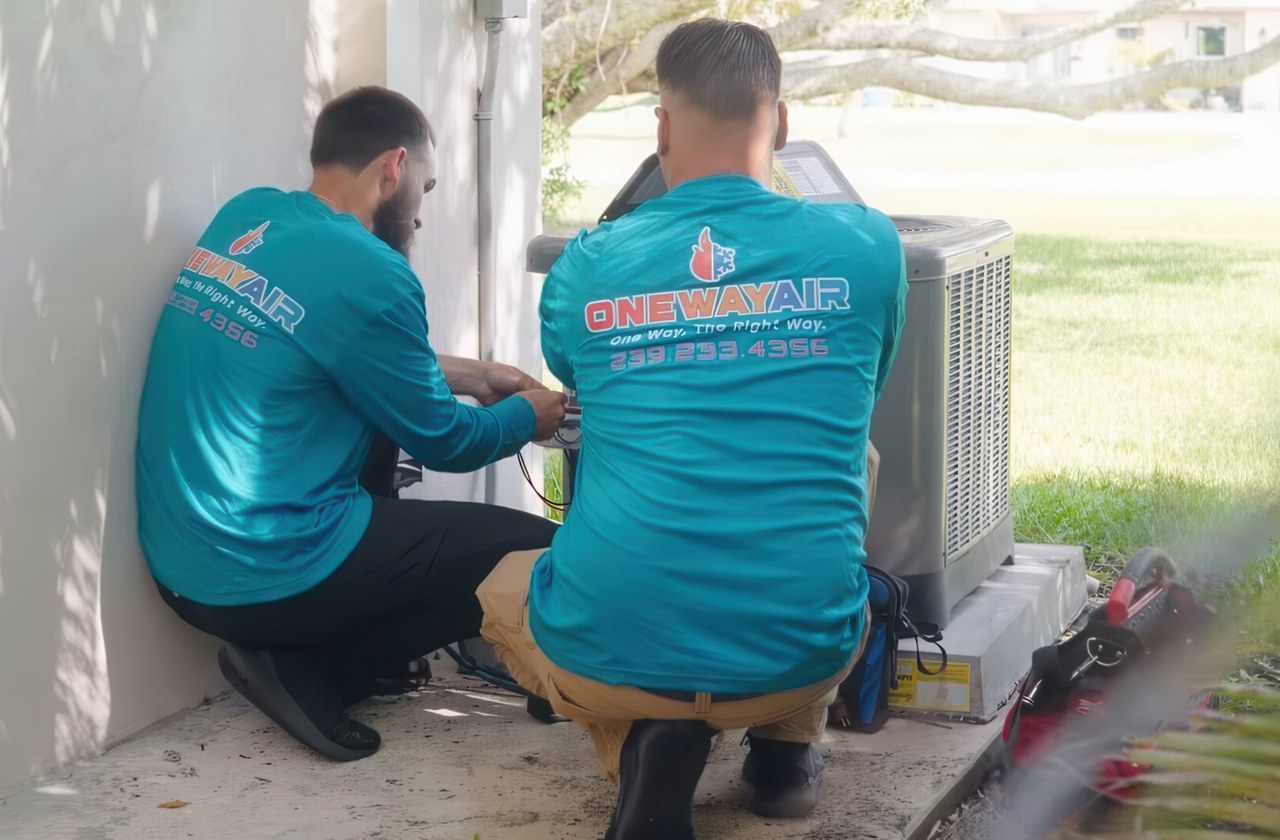Two HVAC technicians in matching uniforms from One Way Air performing maintenance on an outdoor AC unit.
