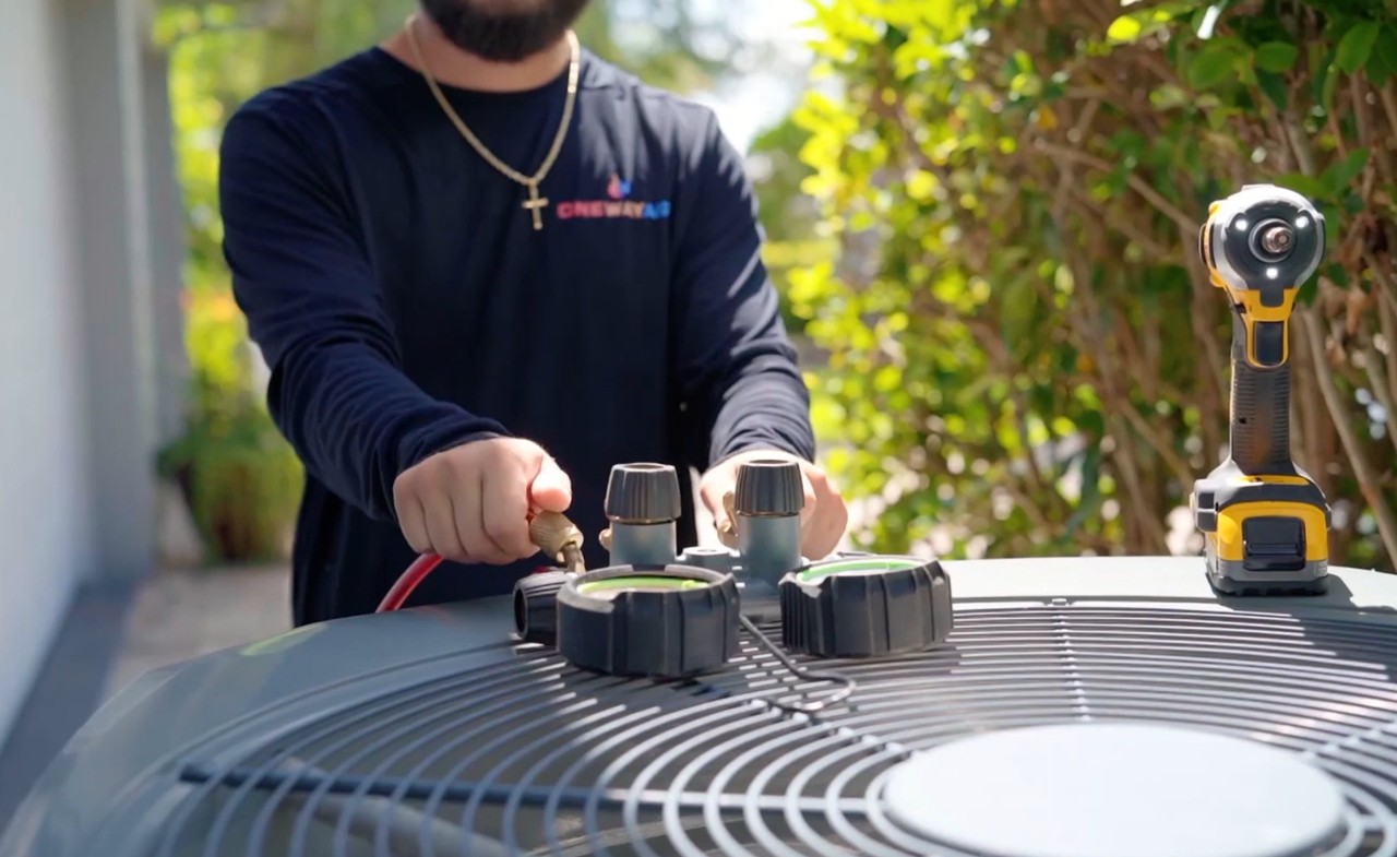 A close-up of an HVAC technician's hands connecting gauges to an outdoor air conditioning unit during maintenance. The technician wears a black long-sleeve shirt with a logo that reads “ONEWAY AIR” and a gold cross necklace. A cordless drill is placed on top of the AC unit, and green foliage is visible in the background.
