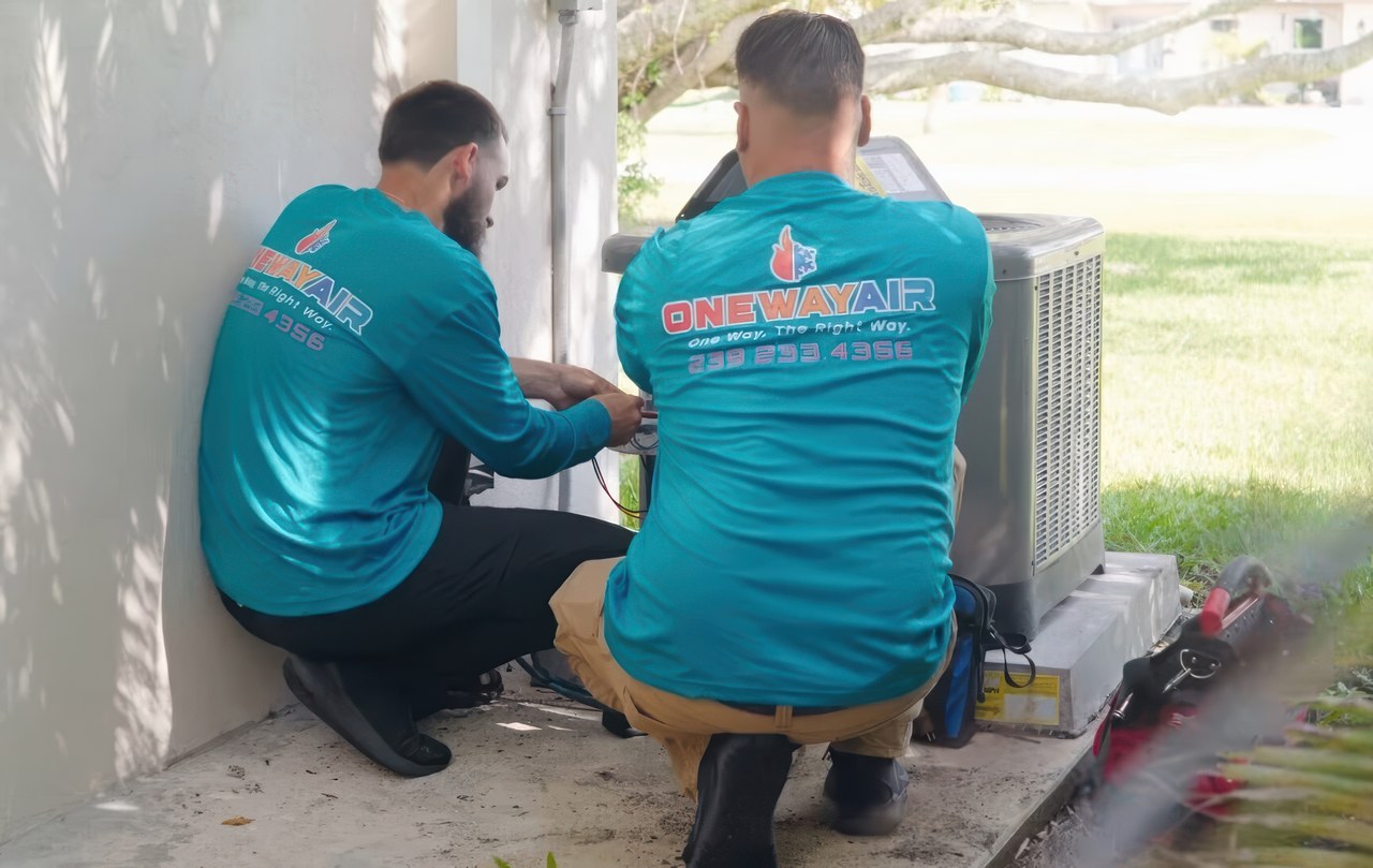 Two HVAC technicians from One Way Air, wearing matching blue uniforms, service an outdoor AC unit on a concrete pad beside a house.