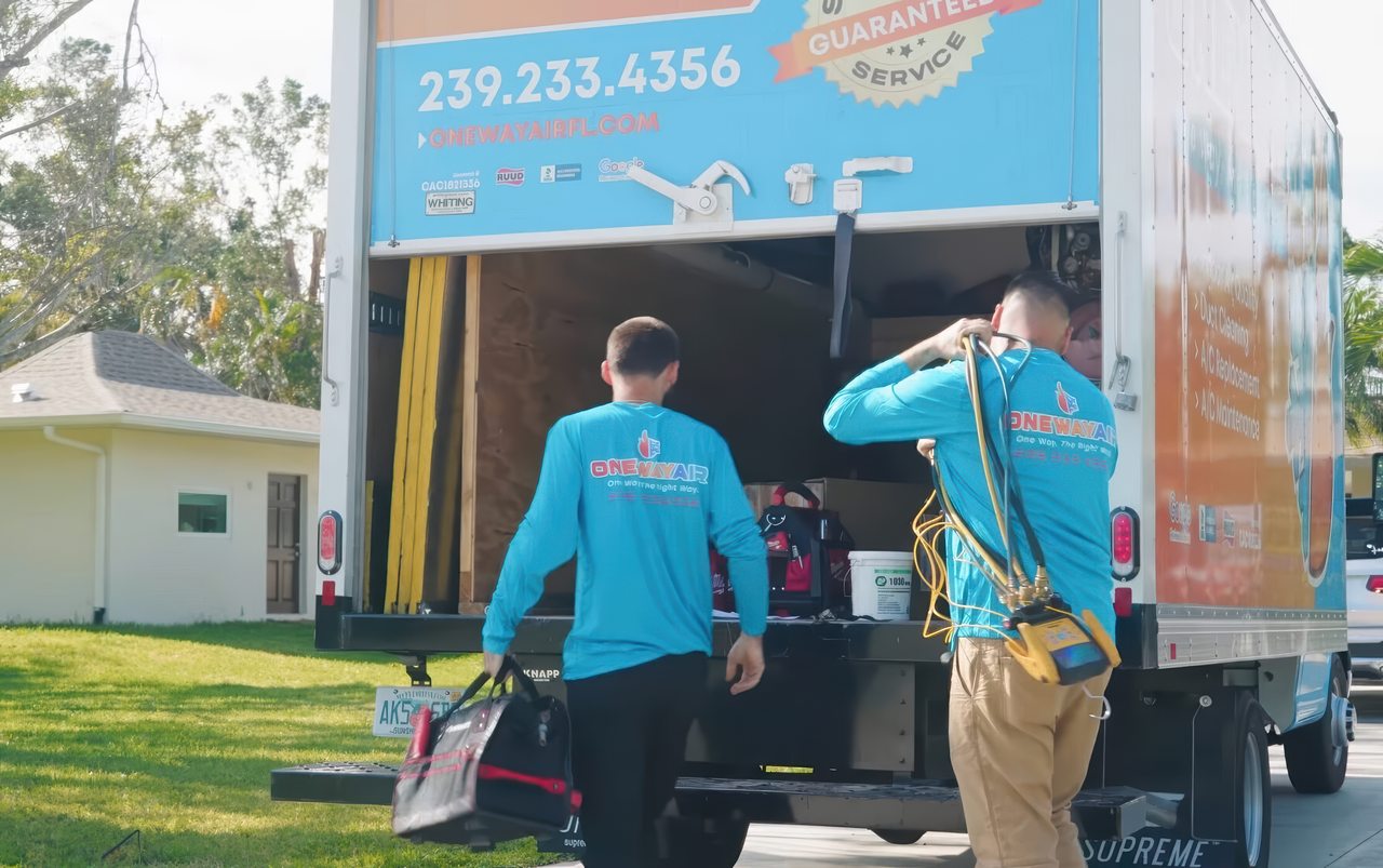 Two One Way Air HVAC technicians unload tools from a service truck on a sunny residential driveway.