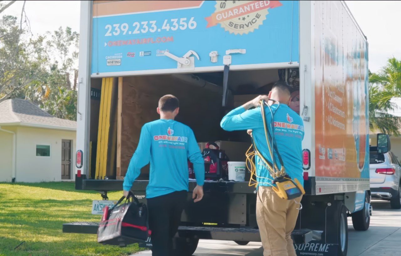 Two service workers in One Way Air uniforms retrieve equipment from a branded truck parked near a residential home under bright daylight.